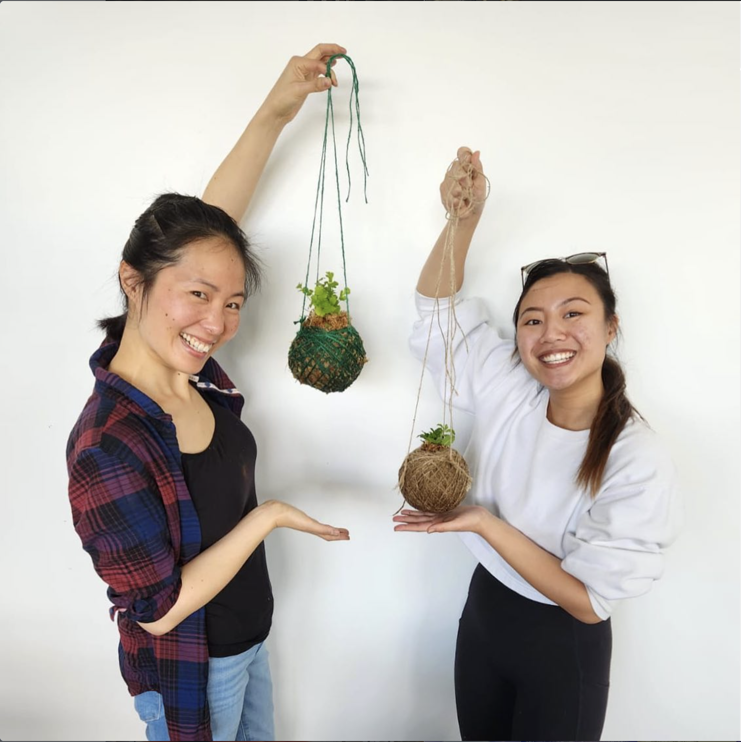Two smiling makers holding their handmade hanging kokedama plant crafts