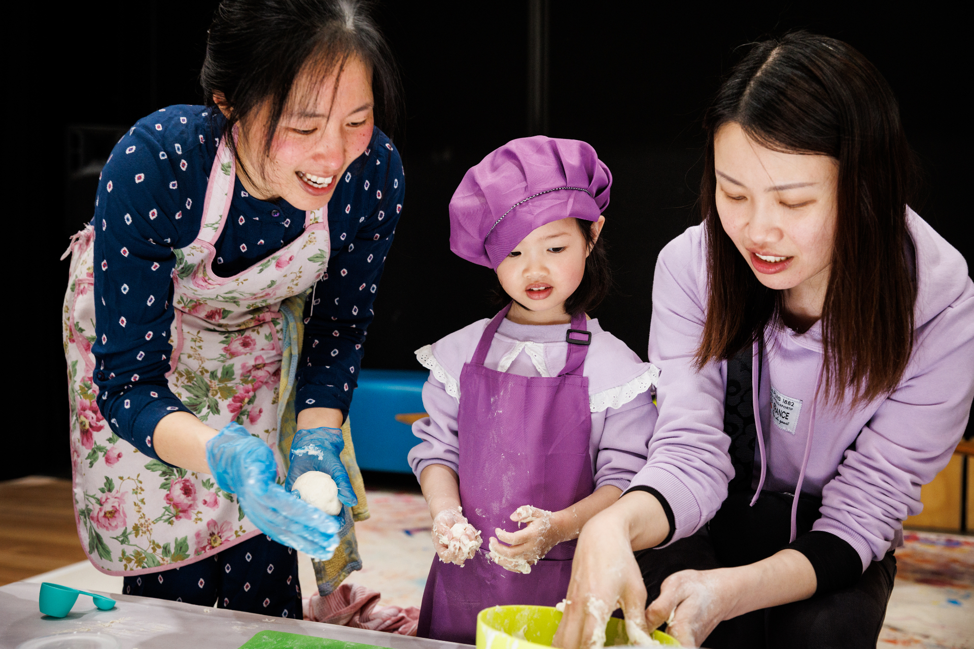 A young chef in a purple apron shaping dough alongside two adults at a family cooking workshop